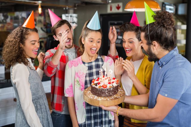 Group of friends celebrating a birthday in a restaurant, with one holding a cake with lit candles. Everyone is wearing party hats and expressing joy and excitement. Perfect for use in advertisements, social media posts, and articles about celebrations, birthdays, and social gatherings.