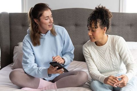 Friends sharing tablet and planning together on cozy bed, casual home collaboration scene