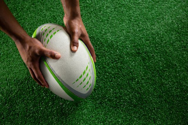 High angle view of hands holding rugby ball on green field. Ideal for use in sports-related content, fitness blogs, team-building activities, and promotional materials for rugby events or training programs.