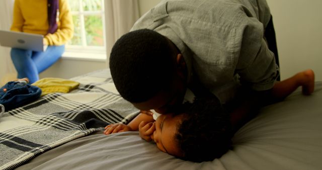 Father is affectionately kissing his child on a bed, creating a tender family moment. A woman in the background is using a laptop, indicating a modern and balanced family lifestyle. Perfect for illustrating concepts of parental love, bonding, family life, modern parenting, and home environments.