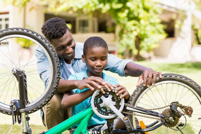 Father and son are repairing a bicycle together outdoors on a sunny day. They are smiling and enjoying the bonding experience. This image can be used for themes related to family activities, parent-child relationships, outdoor fun, and learning new skills.