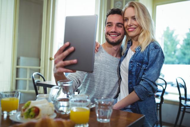 Young couple taking selfie with tablet, sharing joy and capturing moments over breakfast in cozy café. Ideal for concepts of modern lifestyle, digital interaction, social media, and romantic relationships.