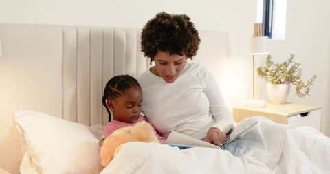Mother reading bedtime story with daughter in cozy daylight bedroom, cuddling teddy bear