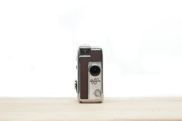 Vintage Revere 40 movie camera placed on a wooden surface against a white background. Ideal for illustrating topics about retro photography, collectibles, and antique film equipment. Suitable for use in articles on the history of cameras, vintage filmmaking, or nostalgic decorative items.