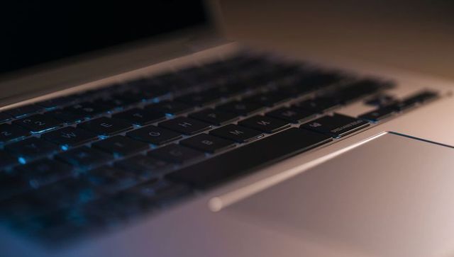 Backlit laptop keyboard and aluminum trackpad glowing with cyan light in dim workspace