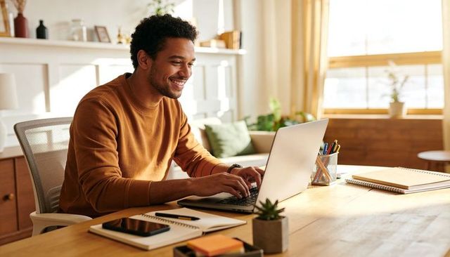 Smiling man working on laptop at sunlit wooden desk with notebooks and plant