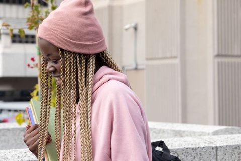 African american teen student wearing pink beanie holding colorful folders on urban campus