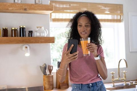 African american woman checking smartphone while holding glass of orange juice in modern kitchen
