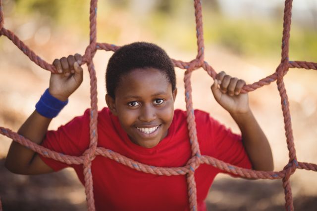 Young boy smiling while leaning on a net during an obstacle course in a boot camp. Ideal for use in advertisements for children's outdoor activities, fitness programs, summer camps, and sports events. Perfect for promoting healthy and active lifestyles for kids.