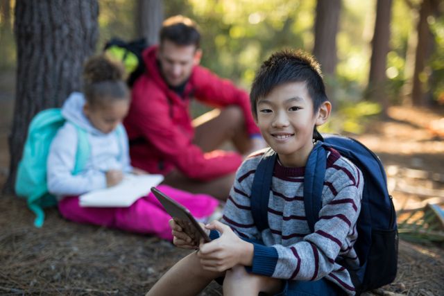 Boy sitting in forest using digital tablet, smiling at camera. Background shows girl writing in notebook and adult supervising. Ideal for themes of outdoor education, family bonding, technology in nature, and childhood adventures.