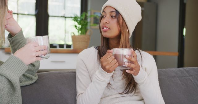 Happy diverse teenager girls sitting on sofa, drinking cocoa and talking. Spending quality time, lifestyle, friendship and adolescence concept.