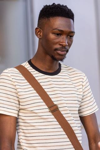 Thoughtful african man standing wearing striped tee and brown crossbody strap, minimalist portrait