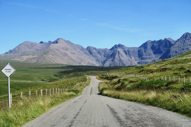 A picturesque road stretches toward majestic mountains under a clear blue sky. Ideal for travel brochures, adventure blogs, and nature photography collections. Promotes outdoor activities, road trips, and tourism in rural settings.