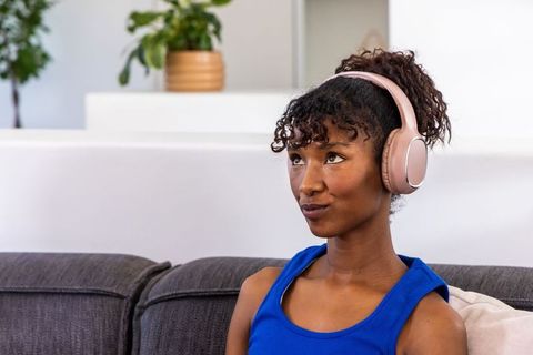 African american woman wearing pink headphones listening and daydreaming on gray couch