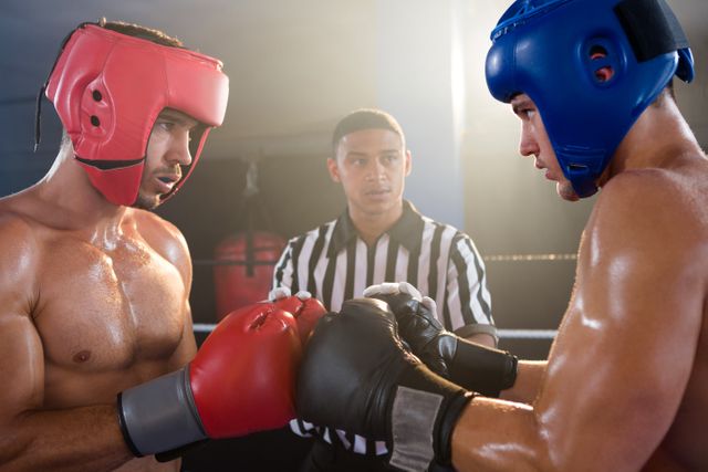 Two male boxers wearing headgear and gloves touching gloves before a match, with a referee observing. This image can be used to depict themes of sportsmanship, competition, and athletic preparation. Ideal for use in sports-related articles, fitness promotions, and advertisements for boxing events.