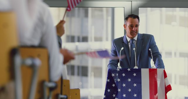 The image shows a politician giving a speech at a campaign rally. The man is standing at a podium decorated with the American flag, addressing an audience holding small American flags. This image is ideal for use in articles or advertisements related to politics, elections, public speaking events, or governmental campaigns.