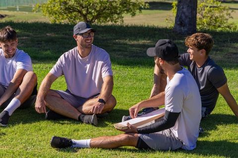 Friends laughing and relaxing on sunny park grass with cricket bat and casual sportswear
