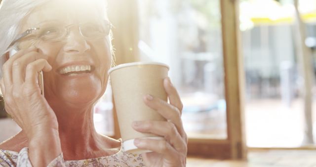 Senior Caucasian woman chats on the phone at a sunny cafe. She enjoys a warm beverage while engaging in a pleasant conversation.