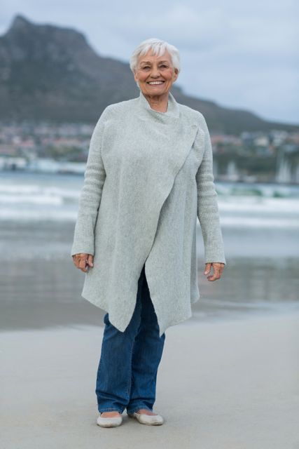 Senior woman with grey hair smiling while standing on a beach with mountains in the background. She is wearing casual clothing including a grey sweater and jeans. Ideal for use in advertisements, articles, or brochures related to retirement, senior lifestyle, health and wellness, travel, and leisure.