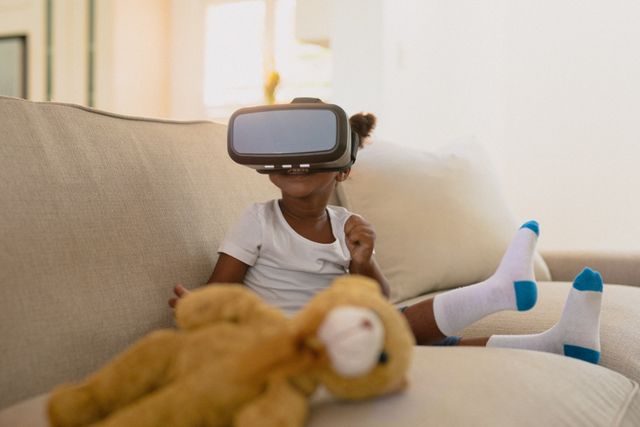 African american girl wearing vr headset and sitting on sofa with teddy bear. family, spending time together at home.