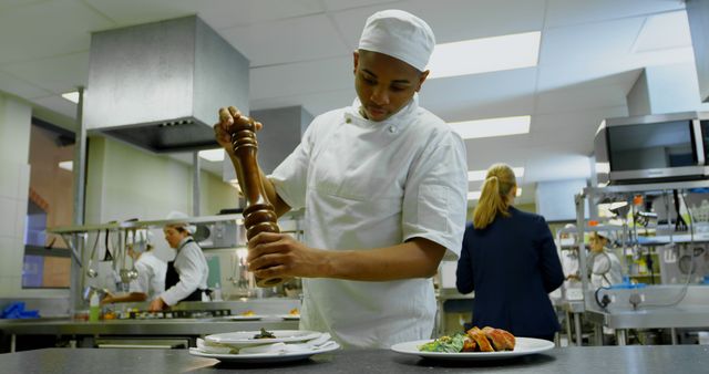 Chef in white uniform using pepper grinder to add seasoning to a plated dish in a modern restaurant kitchen. Other kitchen staff working in background. Perfect for illustrating culinary arts, restaurant advertisements, cooking blogs, and culinary training materials.