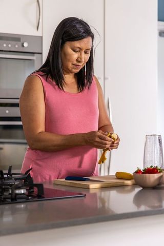 Mature woman peeling banana at kitchen counter preparing healthy smoothie ingredients