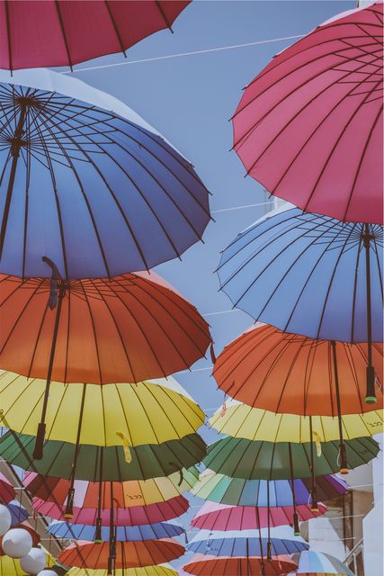 The photo captures multiple colorful umbrellas hanging and creating a vibrant canopy against a clear blue sky. This scene is often seen in art installations or street festivals where colorful umbrellas add a cheerful and whimsical touch. Perfect for use in travel brochures, lifestyle blogs, festival promotions, or decorative art prints.