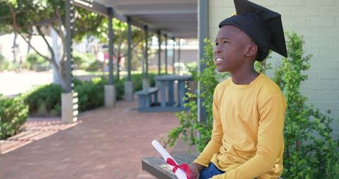 Young african boy wearing graduation cap holding diploma sitting on sunlit campus bench
