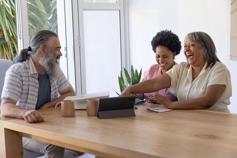 Multigenerational diverse family laughing while using tablet at sunlit wooden dining table
