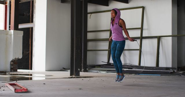 This image shows a woman exercising with a jump rope in an urban building. She wears a pink hoodie and blue pants and is focusing on her workout. Ideal for use in fitness or lifestyle articles, workout guides, athletic gear promotions, and inspirational blogs on fitness and health.