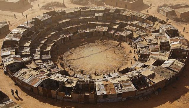 Circular makeshift market ringed by corrugated stalls around cracked desert pit, vendors