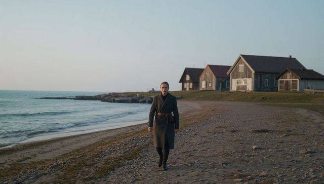 Woman walking along rocky shoreline at golden hour past rustic coastal cottages