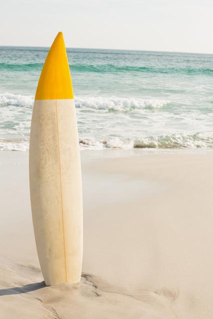 Surfboard standing upright on sandy beach with ocean waves in background. Ideal for use in travel brochures, surfing promotions, summer vacation advertisements, and beach-themed designs.