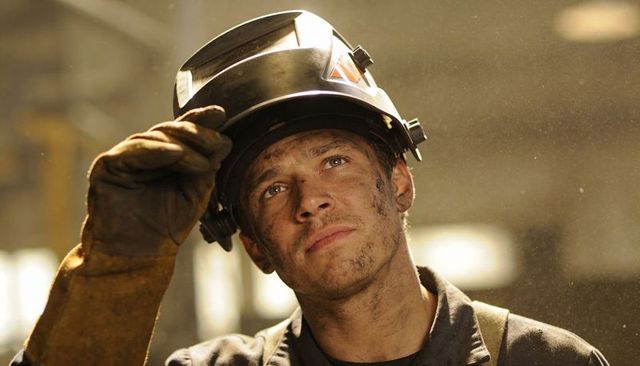 Sooty welder lifting helmet and gazing upward in dusty industrial workshop with warm light