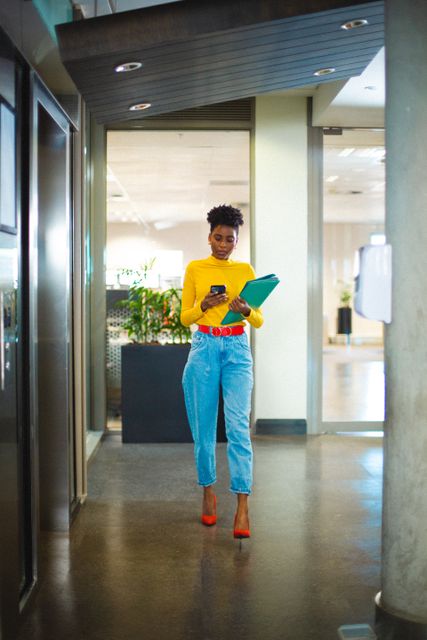 Young African American businesswoman walking through a modern office while using a smartphone and holding files. Ideal for use in business, technology, and corporate settings to depict professionalism, modern work environments, and productivity.