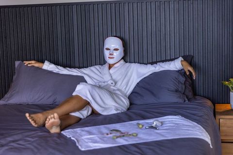 African-american woman relaxing in robe with led facial mask on bed for skincare wellness