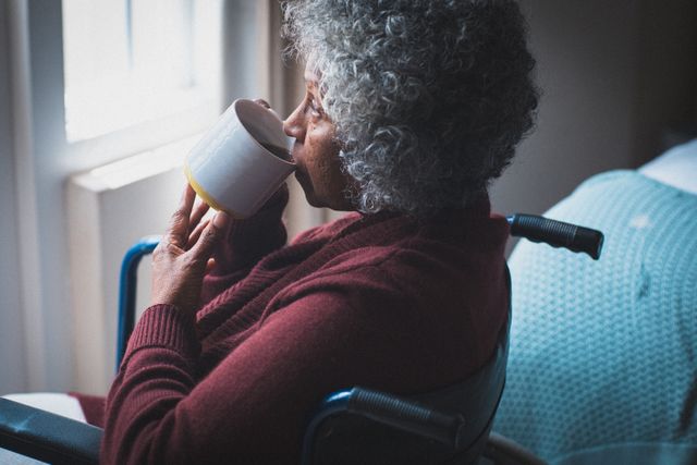 This image depicts a senior African American woman in a wheelchair, thoughtfully drinking tea while looking out of a window. The natural light and cozy setting create a peaceful and reflective atmosphere. This image can be used in articles or advertisements related to elderly care, home living, mental health, isolation during quarantine, or morning routines.