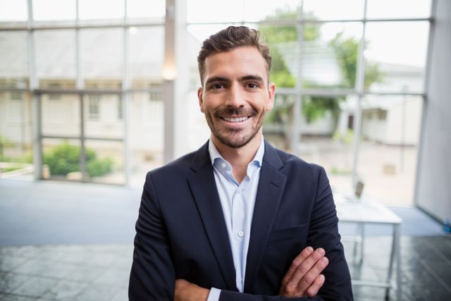 This image features a confident businessman smiling with arms crossed in a modern office environment. Ideal for use in corporate websites, business presentations, leadership articles, and professional networking profiles.