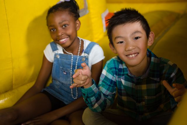 Two happy children, a girl and a boy, are sitting on a bouncy castle, smiling and enjoying their time. The girl is wearing a white shirt and denim overalls, while the boy is in a green plaid shirt. This image can be used for promoting children's events, indoor playgrounds, summer camps, or any content related to childhood fun and friendship.