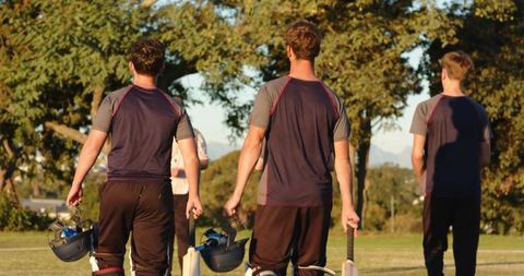 Diverse male cricket players walking to coach on golden field tapping bats carrying helmets