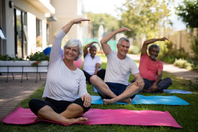 Senior individuals are sitting on yoga mats in a park, engaging in a group exercise session. They are stretching with hands raised, promoting physical health and wellness. This image is ideal for use in health and wellness campaigns, senior fitness programs, community activity promotions, and advertisements for outdoor exercise equipment.