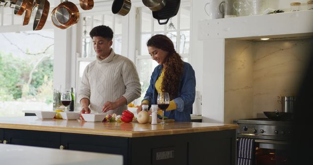 Young couple is preparing a meal together in a stylish, modern kitchen. They are chopping vegetables and smiling, creating a warm, collaborative atmosphere. Wine glasses and fresh ingredients are on the counter. Perfect for advertisements related to home life, cooking, kitchen appliances, or couple activities.