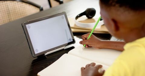 African american boy studying at home using tablet and writing in notebook