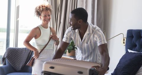 Multiracial couple packing luggage in sunlit modern hotel bedroom preparing for trip