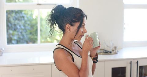 Woman sipping hot drink in sunlit minimalist kitchen holding green mug