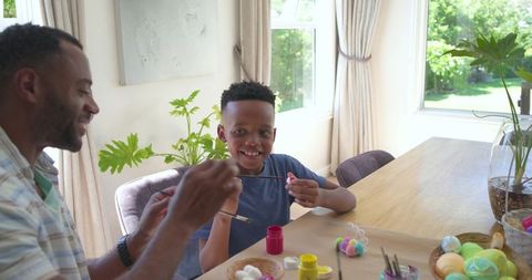 African american father and son painting easter eggs together at home dining table