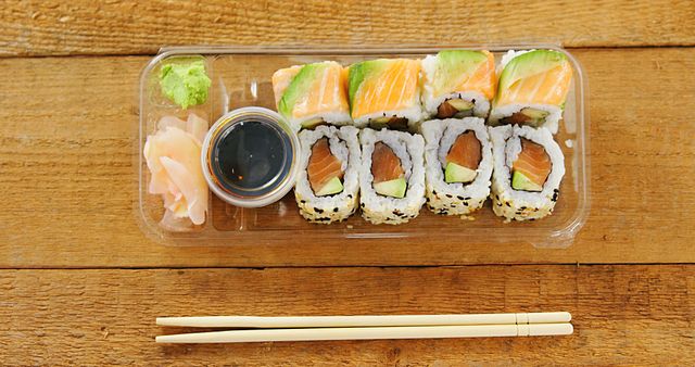 A variety of sushi rolls with salmon and avocado are neatly arranged in a plastic tray, complete with soy sauce, wasabi, and pickled ginger, with copy space. Chopsticks rest beside the tray, ready for a delicious Japanese cuisine experience.