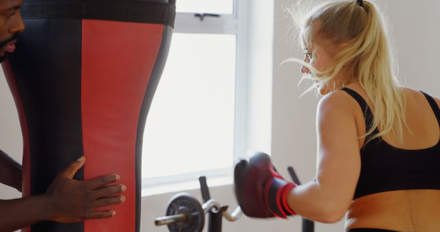 A young Caucasian woman is practicing boxing, punching a heavy bag held by an African American man, with copy space. Her focus and determination are evident as she engages in this intense fitness training session.