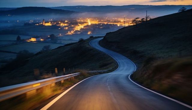Winding country road descending into lit valley at dusk with mist, guardrail, headlights