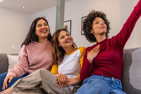 Multicultural women laughing and taking selfie on couch, cozy friends holding hands
